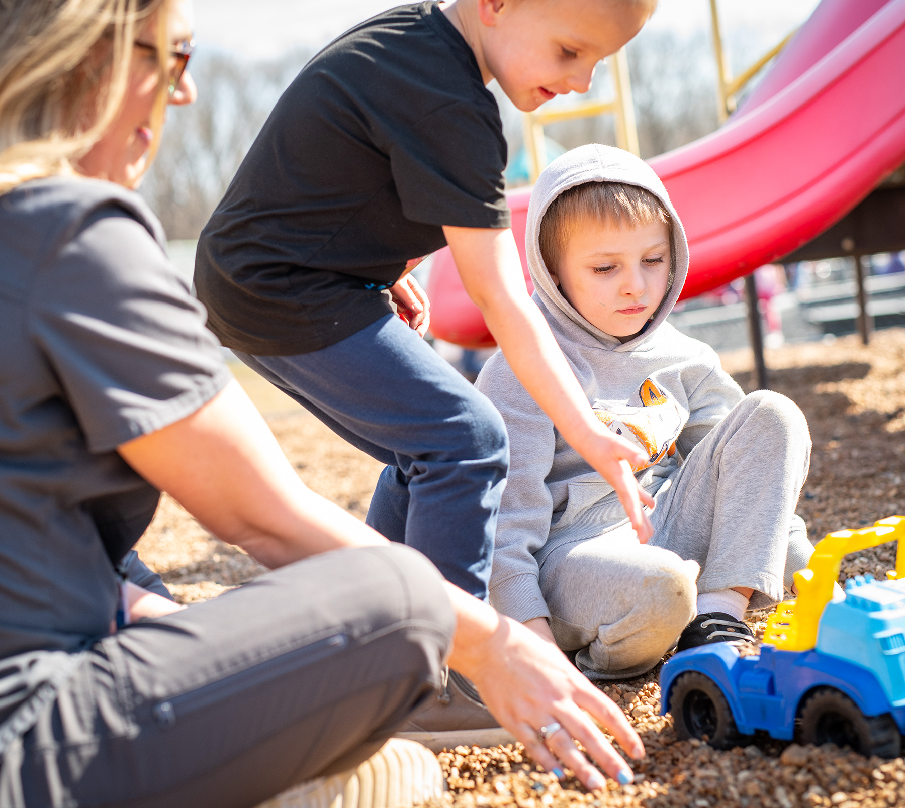 Photo of two children playing with trucks and sand in a playground, supervised by an adult
