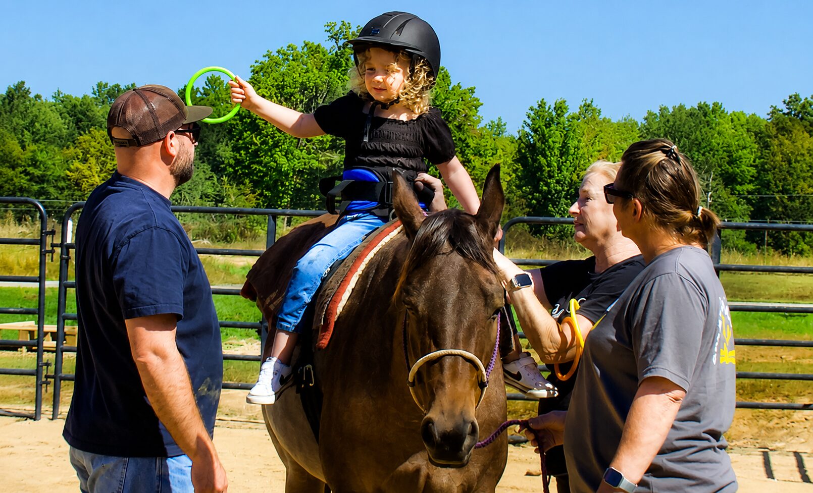 A child on a horse handing a plastic ring to a therapist standing next to them